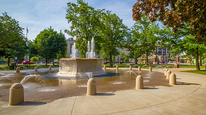 A campus with a large fountain surrounded by smaller fountains in a circle with trees in the background.