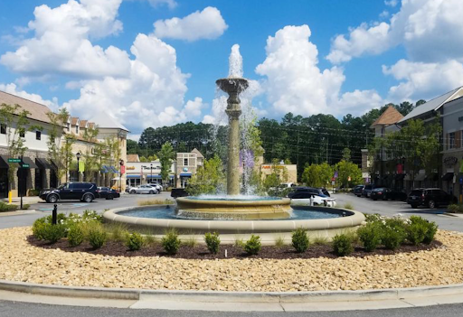 Two-tier classical fountain in a landscaped roundabout at a retail center, positioned between parking and storefronts.